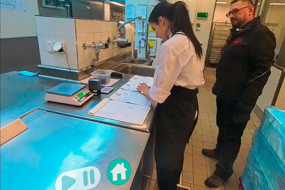 Durch die Ansicht der VR-Brille sieht man eine Auszubildende Köchin und ihren Berufsbildner, wie sie in der Küche etwas lesen.