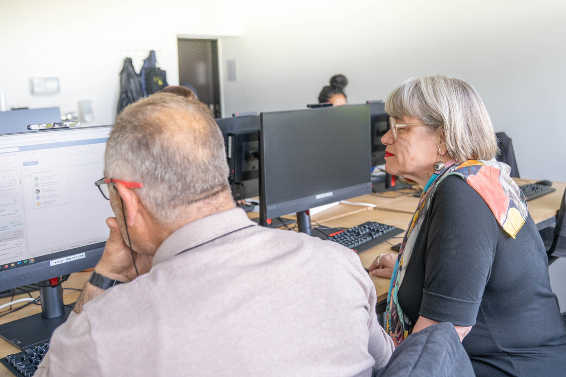 die Kursleiterin sitzt neben einem Kursteilnehmer und unterstützt ihn bei einer Übung am Computer