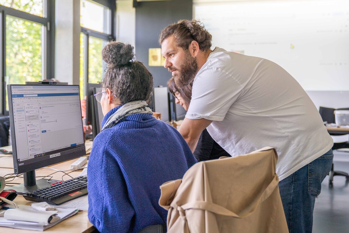 ein RAV-Personalberater schaut einer Kursteilnehmerin bei einer Übung am Computer über die Schulter und hilft ihr beim Lösen der Aufgabe