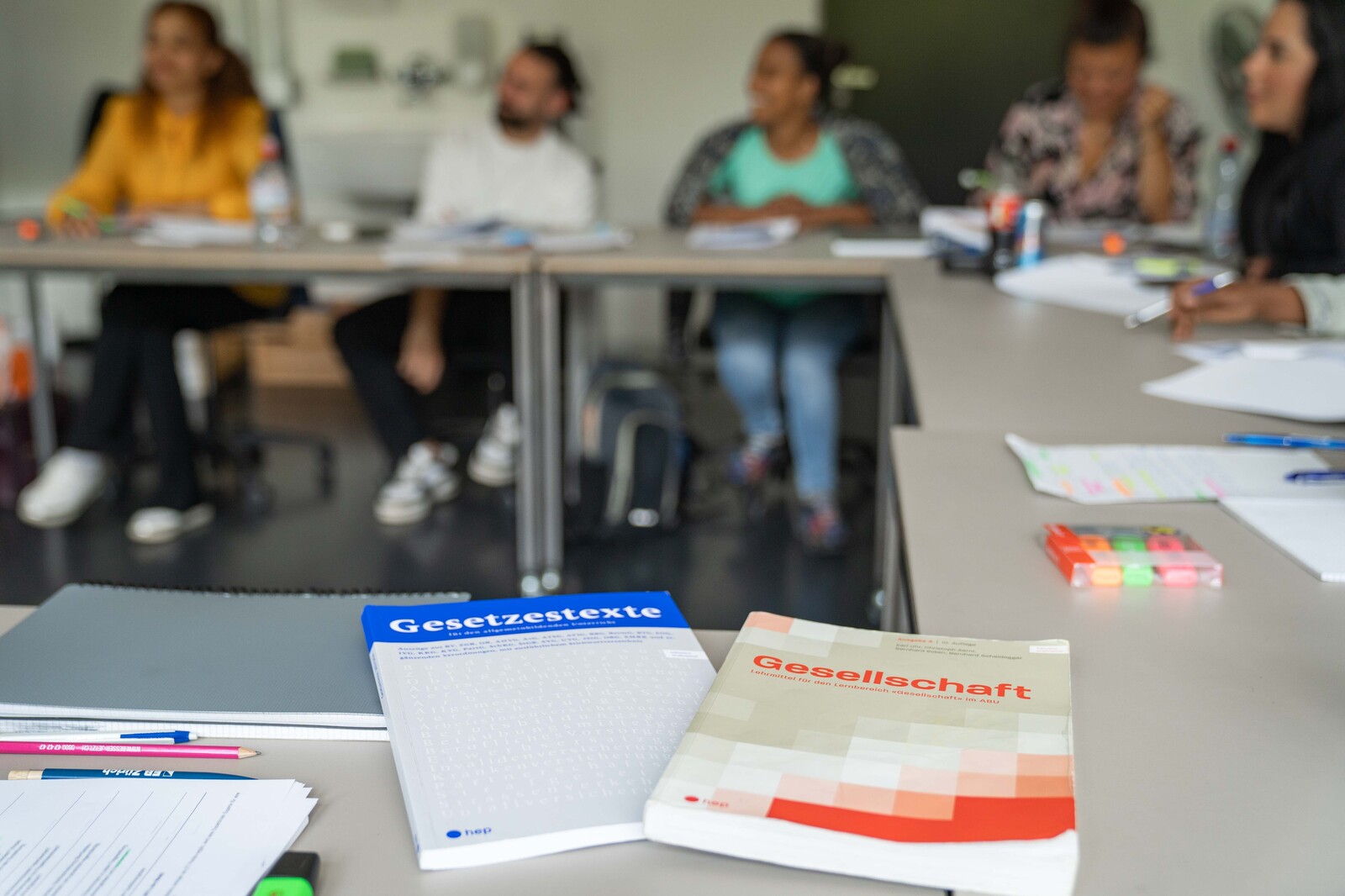Zwei Bücher aus einem ABU-Kurs liegen auf einen Tisch, im Hintergrund sieht man mehrere Kursteilnehmer